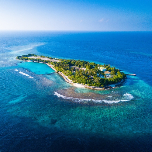 Aerial view of a small, lush tropical island surrounded by clear turquoise waters and gentle waves. The island features dense greenery and a few structures, evoking a serene, isolated paradise.