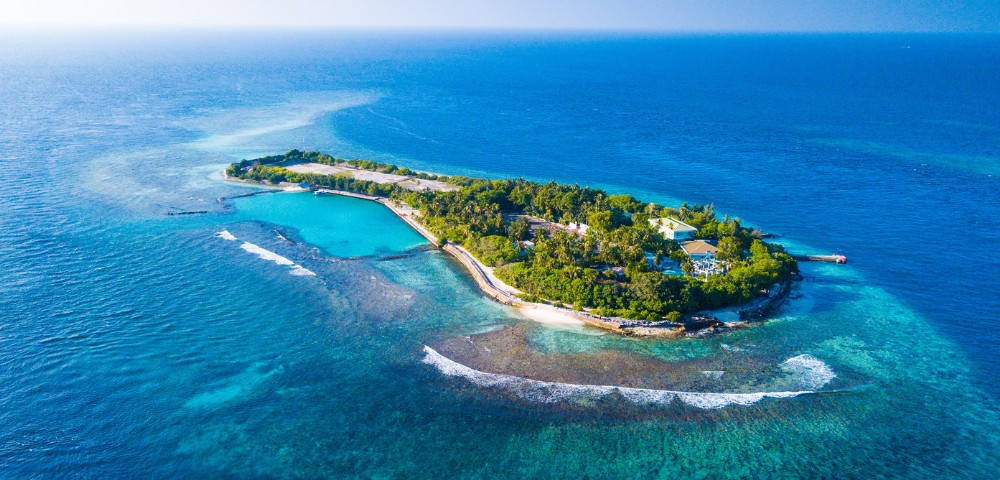 Aerial view of a small, lush tropical island surrounded by clear turquoise waters and gentle waves. The island features dense greenery and a few structures, evoking a serene, isolated paradise.