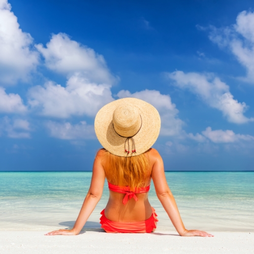 A woman in a straw hat sits on a white sandy beach, facing a tranquil turquoise sea and blue sky with fluffy clouds, evoking a sense of relaxation.