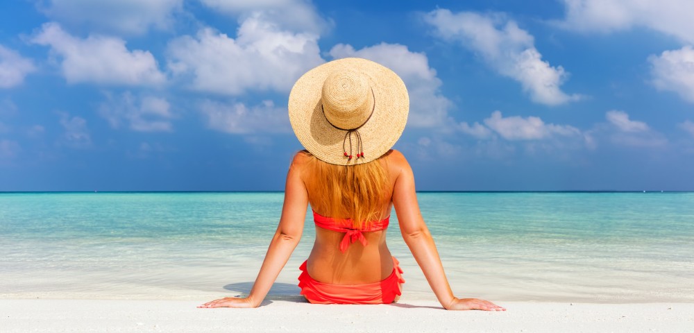A woman in a straw hat sits on a white sandy beach, facing a tranquil turquoise sea and blue sky with fluffy clouds, evoking a sense of relaxation.