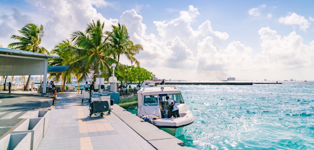 A sunny coastal scene with a white boat docked at a pier, surrounded by turquoise water and lush palm trees. The sky is bright with fluffy clouds.