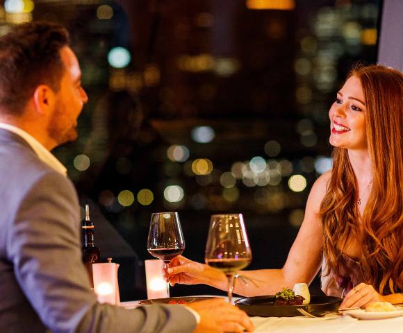 Two people enjoying drinks at a restaurant table with blurred city lights in the background.