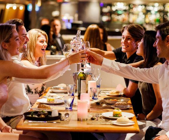 Group of people clinking glasses over a meal in a lively restaurant setting.