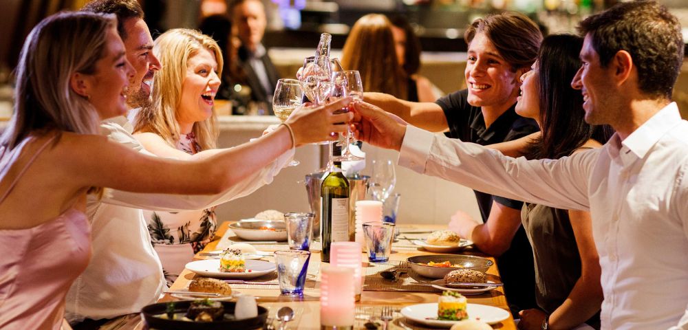 Group of people clinking glasses over a meal in a lively restaurant setting.