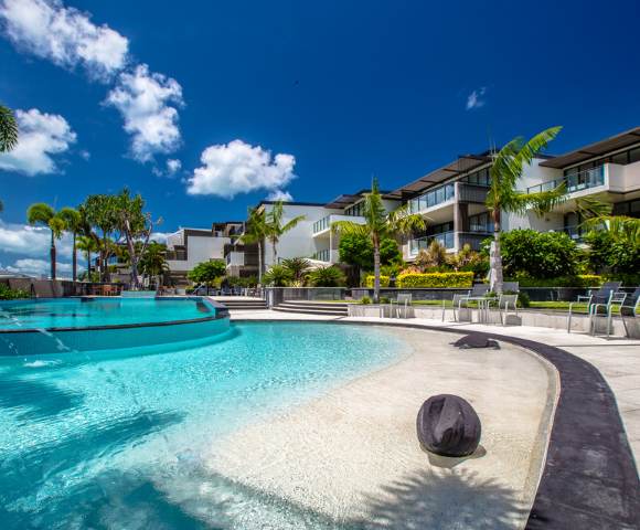 Wide view of a resort pool area surrounded by lush greenery under sunny skies.