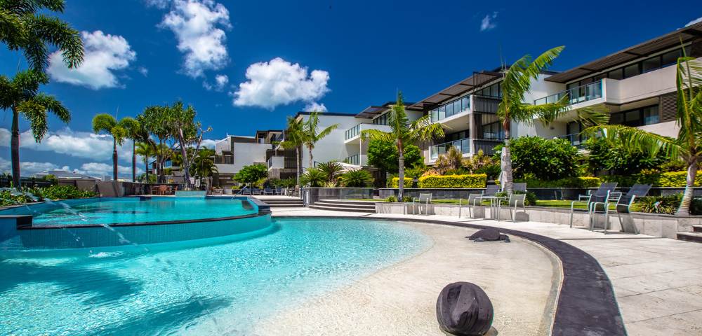 Wide view of a resort pool area surrounded by lush greenery under sunny skies.