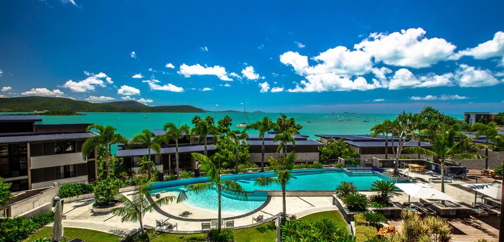 Elevated view of a resort pool area with surrounding greenery and distant ocean views.