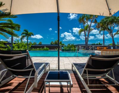 Two lounge chairs facing an outdoor pool with palm trees and blue skies in the background.