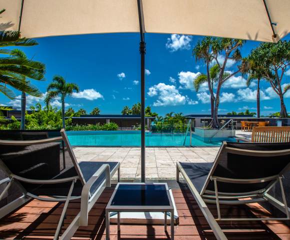 Two lounge chairs facing an outdoor pool with palm trees and blue skies in the background.