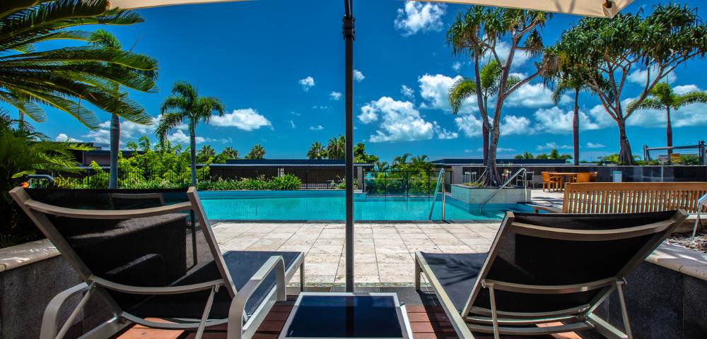 Two lounge chairs facing an outdoor pool with palm trees and blue skies in the background.