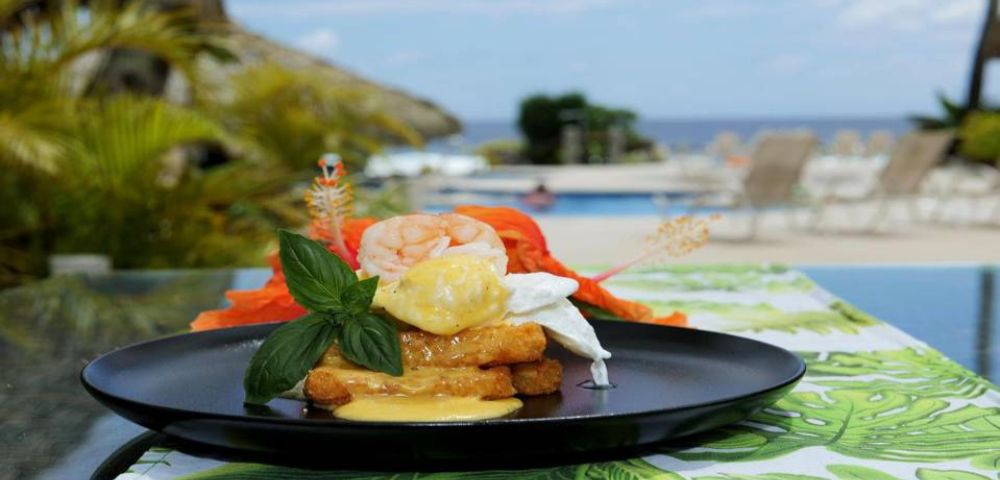 Gourmet dish featuring shrimp, poached eggs, and herbs on a black plate, with a tropical poolside backdrop and vibrant garnish.