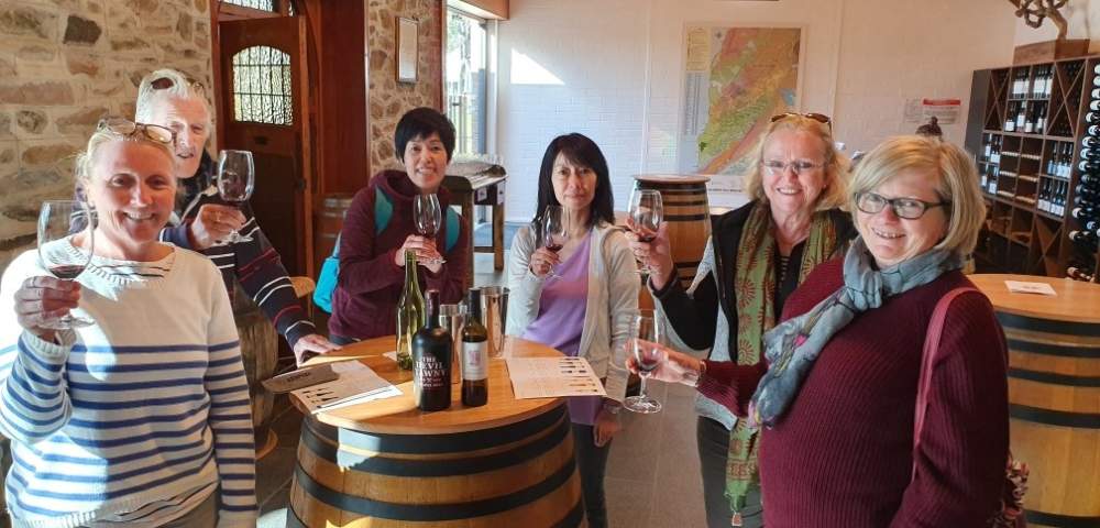 Two people smiling at the camera while standing at a table with wine glasses and bottles.