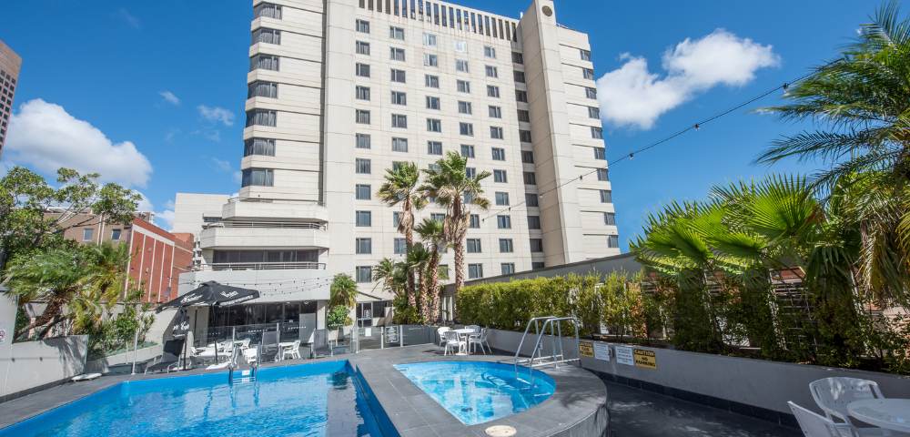 Outdoor pool area at Hotel Grand Chancellor Adelaide surrounded by lounge chairs, palm trees, and greenery.