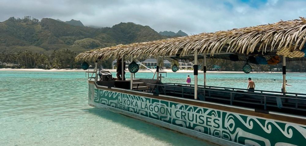 A colorful lagoon cruise boat with a thatched roof docked in clear blue waters, surrounded by mountains and lush tropical scenery.