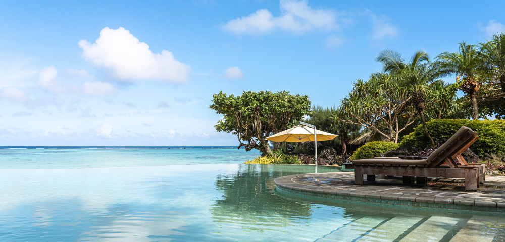 The image features a large, blue swimming pool with a beautiful view of the ocean in the background. The pool is surrounded by a patio area with a bench and a dining table, providing a relaxing spot for people to enjoy the ocean view. There are several umbrellas placed around the patio, offering shade and protection from the sun. In the distance, a few boats can be seen floating on the water, adding to the picturesque scene. The overall atmosphere of the image is serene and inviting, perfect for a day of leisure and relaxation. --ar 580:480 --q 2 --s 750 --v 5.2