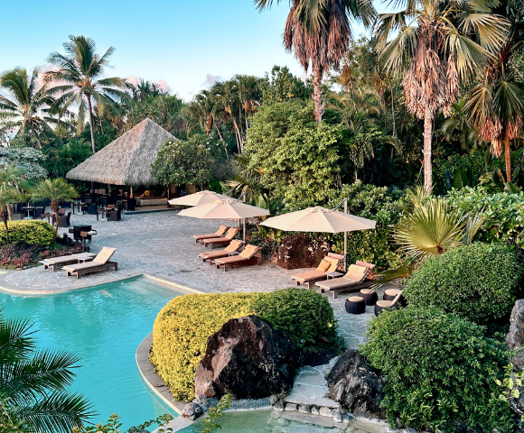 The image features a beautiful tropical resort with a large swimming pool surrounded by lounge chairs and umbrellas. There are several umbrellas providing shade for the guests, with some placed near the pool and others scattered throughout the resort.