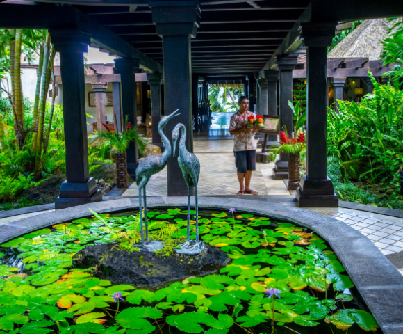 A tranquil resort entrance featuring a stone pond with lotus flowers, and two elegant cranes amidst lush greenery.