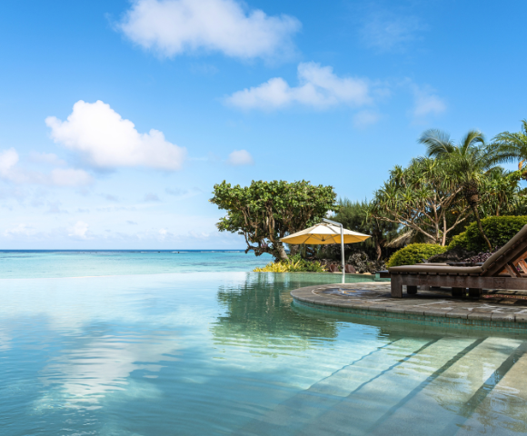 The image features a large, blue swimming pool with a beautiful view of the ocean in the background. The pool is surrounded by a patio area with a bench and a dining table, providing a relaxing spot for people to enjoy the ocean view. There are several umbrellas placed around the patio, offering shade and protection from the sun. In the distance, a few boats can be seen floating on the water, adding to the picturesque scene. The overall atmosphere of the image is serene and inviting, perfect for a day of leisure and relaxation. --ar 580:480 --q 2 --s 750 --v 5.2