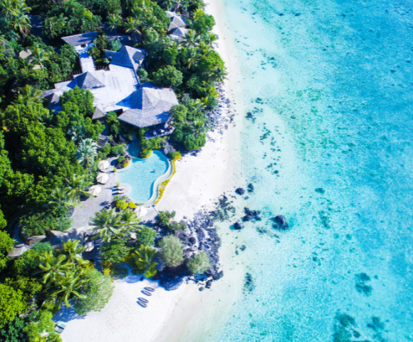 The image features a beautiful beach setting with a house situated on the sandy shore. The house is surrounded by palm trees, creating a tropical atmosphere. In the foreground, there is a large swimming pool, which adds to the luxurious and relaxing ambiance of the scene.