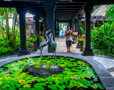 A tranquil resort entrance featuring a stone pond with lotus flowers, and two elegant cranes amidst lush greenery.