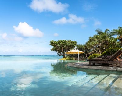 The image features a large, blue swimming pool with a beautiful view of the ocean in the background. The pool is surrounded by a patio area with a bench and a dining table, providing a relaxing spot for people to enjoy the ocean view. There are several umbrellas placed around the patio, offering shade and protection from the sun. In the distance, a few boats can be seen floating on the water, adding to the picturesque scene. The overall atmosphere of the image is serene and inviting, perfect for a day of leisure and relaxation. --ar 580:480 --q 2 --s 750 --v 5.2