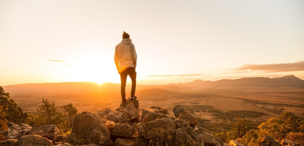 A person in a hoodie stands on rocky terrain, gazing at a glowing sunrise over a vast landscape of fields and distant mountains, evoking serenity and awe.