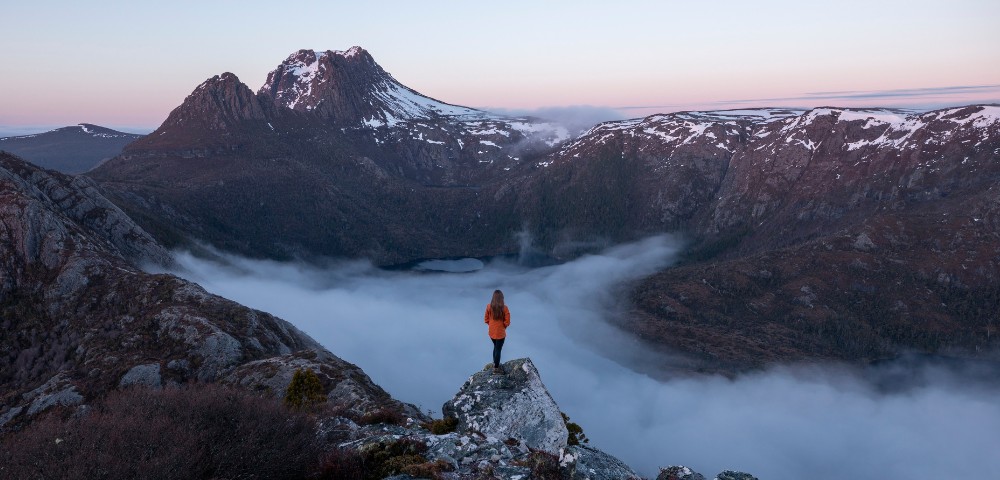 A person in an orange jacket stands on a rock, overlooking a foggy valley with rugged mountains and patches of snow under a pastel pink sky at sunrise.