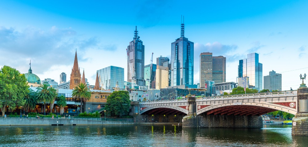 Wide view of a cityscape with modern skyscrapers and historic buildings by a river. A bridge spans the water, surrounded by lush greenery and palm trees.