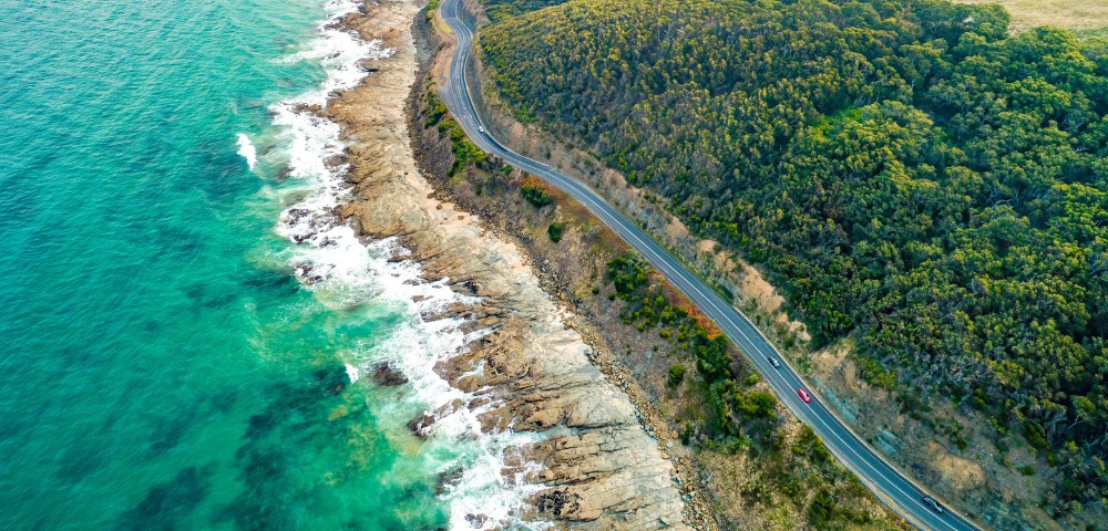 Aerial view of a coastal road winding through lush green hills, with turquoise ocean waves crashing against rocky shorelines on the left. A red car drives along the road, suggesting a serene, scenic journey.