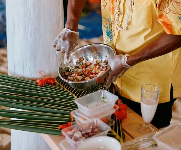 Person wearing gloves and a yellow floral shirt preparing food in a metal bowl, surrounded by plastic containers, a glass of white liquid, and tropical decorations including palm leaves and red flowers.