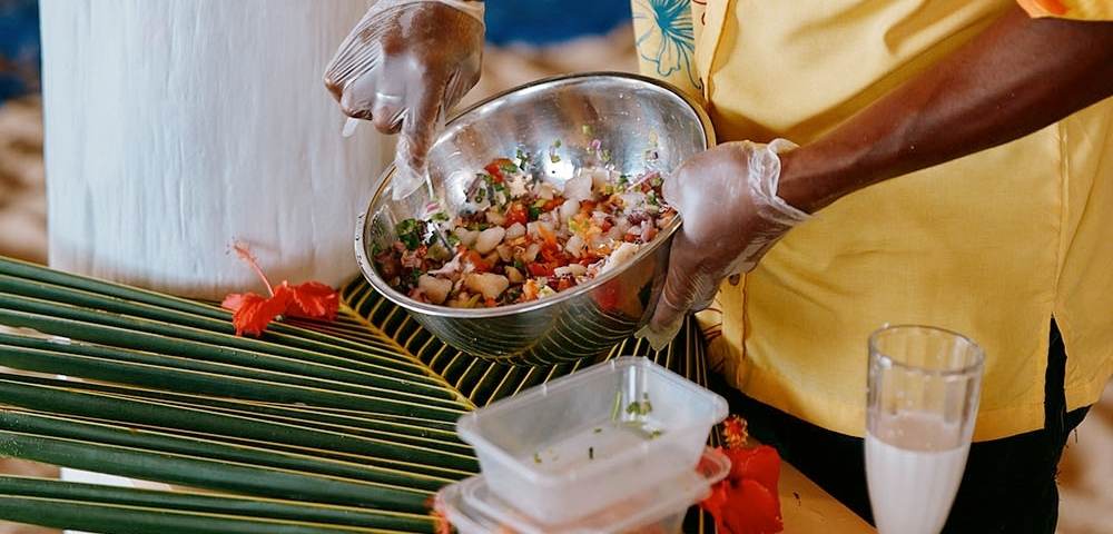 Person wearing gloves and a yellow floral shirt preparing food in a metal bowl, surrounded by plastic containers, a glass of white liquid, and tropical decorations including palm leaves and red flowers.