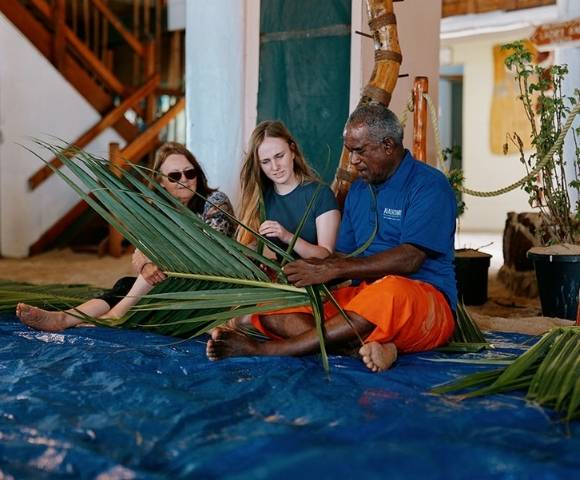 Three individuals seated on a blue tarp indoors, weaving palm fronds together in a setting with potted plants and wooden structures, suggesting a cultural or educational activity.
