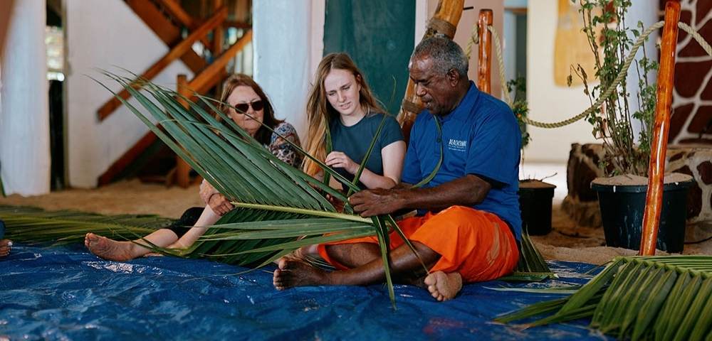 Three individuals seated on a blue tarp indoors, weaving palm fronds together in a setting with potted plants and wooden structures, suggesting a cultural or educational activity.