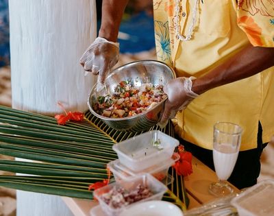 Person wearing gloves and a yellow shirt preparing a colorful salad or ceviche in a metal bowl, with tropical decorations including palm leaves and red flowers on the table.
