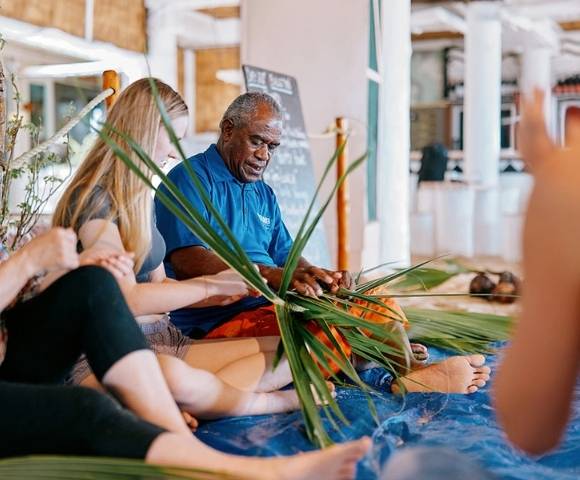 Group of people seated indoors, participating in a hands-on weaving activity using green palm leaves, with one person demonstrating the technique in a communal or cultural setting
