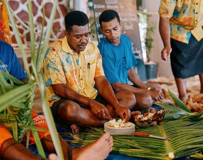 Group of people seated indoors on a mat made of green leaves, preparing and sharing food placed on plates, dressed in colorful clothing with decorative elements in the background