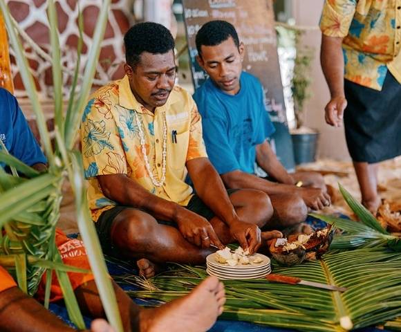 Group of people seated indoors on a mat made of green leaves, preparing and sharing food placed on plates, dressed in colorful clothing with decorative elements in the background