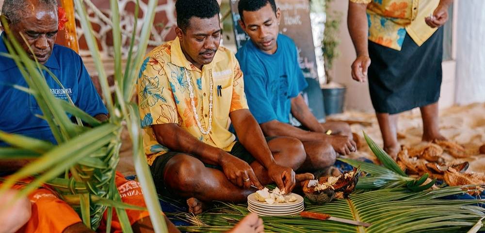 Group of people seated indoors on a mat made of green leaves, preparing and sharing food placed on plates, dressed in colorful clothing with decorative elements in the background
