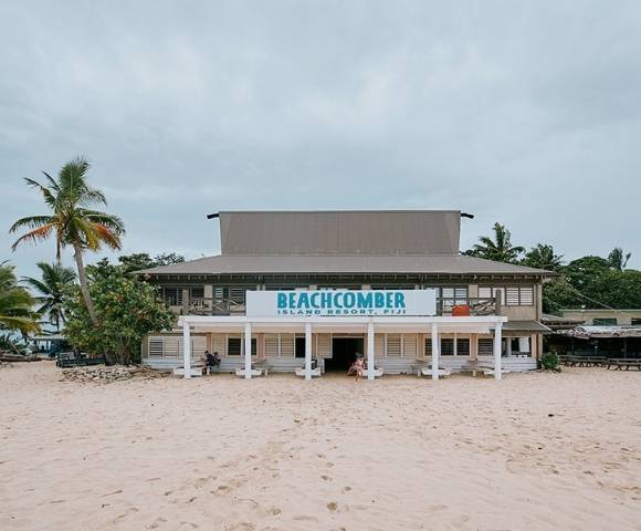 Beachfront building with a sign reading 'BEACHCOMBER ISLAND RESORT FIJI,' surrounded by palm trees and sand, under an overcast sky.