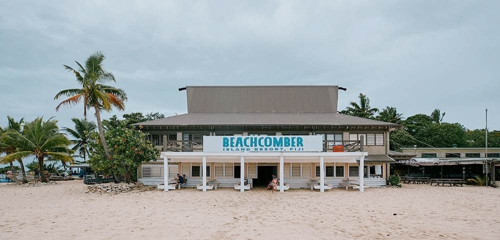 Beachfront building with a sign reading 'BEACHCOMBER ISLAND RESORT FIJI,' surrounded by palm trees and sand, under an overcast sky.