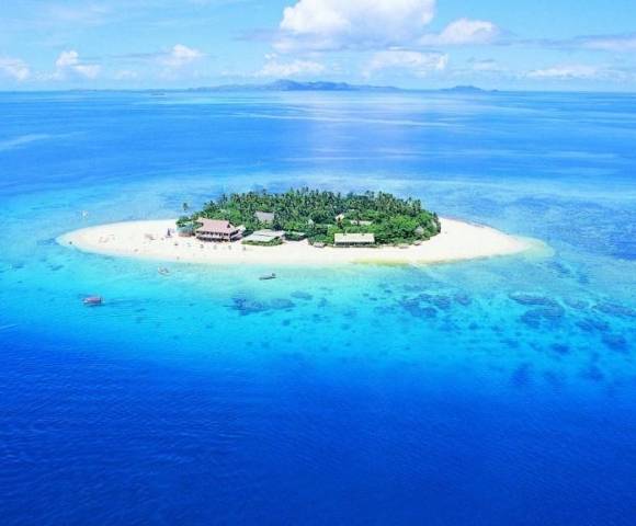Aerial view of a small tropical island with lush greenery, resort buildings, and surrounding clear blue waters with visible coral reefs and sandbanks under a mostly clear sky.