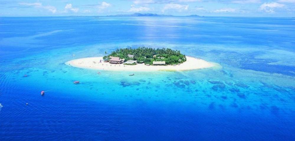 Aerial view of a small tropical island with lush greenery, resort buildings, and surrounding clear blue waters with visible coral reefs and sandbanks under a mostly clear sky.