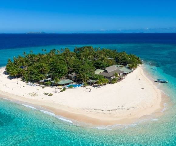 A small tropical island with lush greenery, sandy beaches, resort buildings, and a boat docked along the shoreline, surrounded by clear turquoise waters.