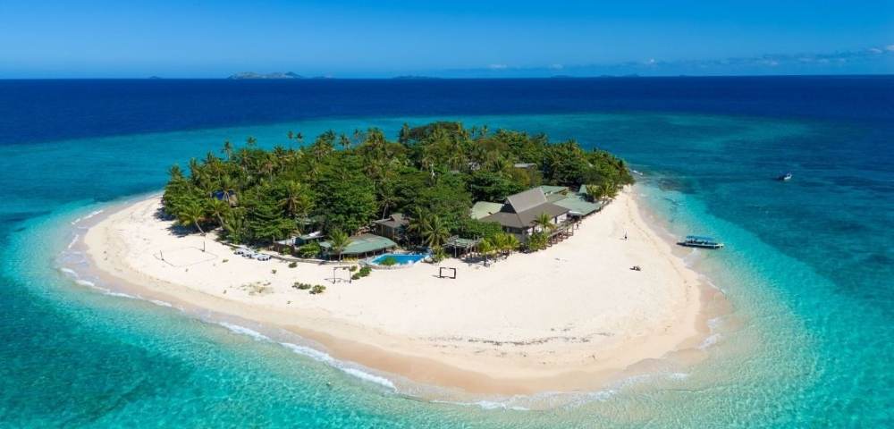 A small tropical island with lush greenery, sandy beaches, resort buildings, and a boat docked along the shoreline, surrounded by clear turquoise waters.