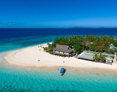Aerial view of a small tropical island with white sandy beaches, lush greenery, several thatched-roof buildings, and a boat anchored near the shore in clear turquoise water