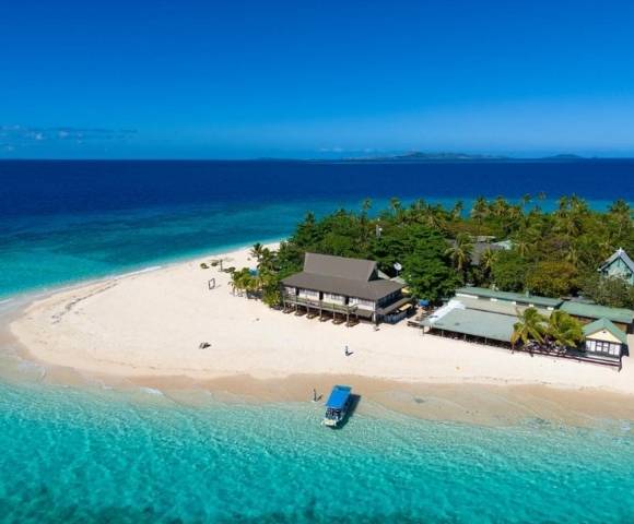 Aerial view of a small tropical island with white sandy beaches, lush greenery, several thatched-roof buildings, and a boat anchored near the shore in clear turquoise water