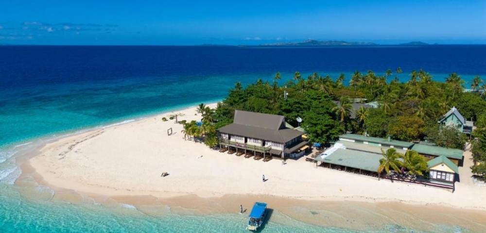 Aerial view of a small tropical island with white sandy beaches, lush greenery, several thatched-roof buildings, and a boat anchored near the shore in clear turquoise water