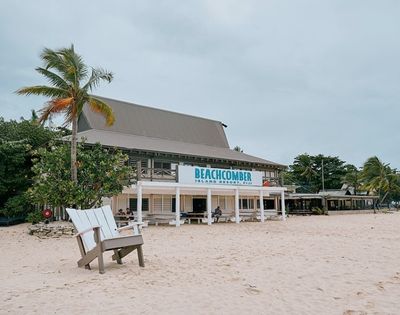 Beachfront view of Beachcomber Island Resort in Fiji, featuring a thatched-roof building with a sign, surrounded by palm trees and a wooden chair on the sandy beach under an overcast sky