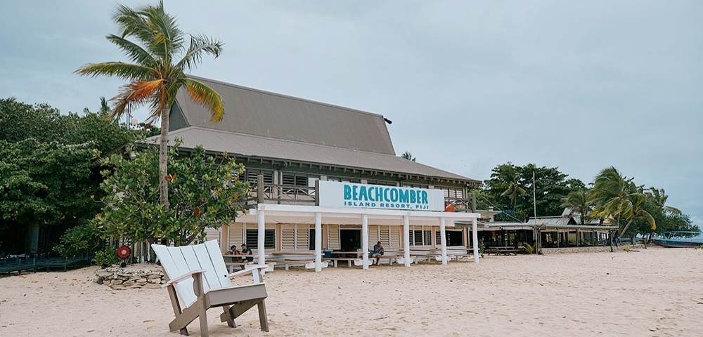 Beachfront view of Beachcomber Island Resort in Fiji, featuring a thatched-roof building with a sign, surrounded by palm trees and a wooden chair on the sandy beach under an overcast sky