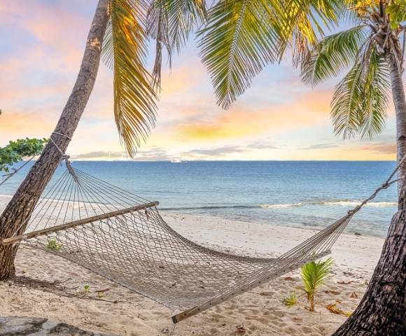 Peaceful beach scene with an empty hammock strung between two palm trees on sandy shore, facing a calm ocean under a colorful sunrise or sunset sky.